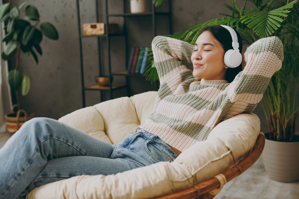 Woman wearing headphones and a cosy sweater lounges back in an arm chair, arm's closed and smiling