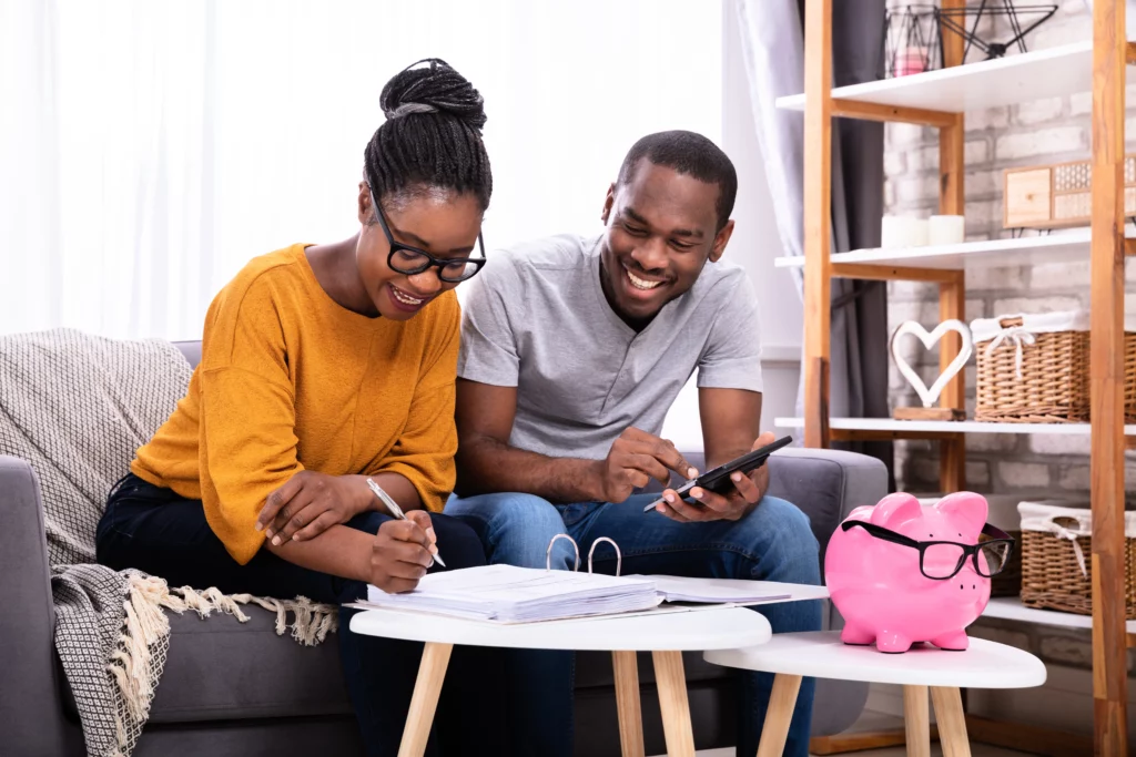 A smiling couple make notes in a binder to hit their saving goals. In front of them sits a pink piggy bank wearing glasses.