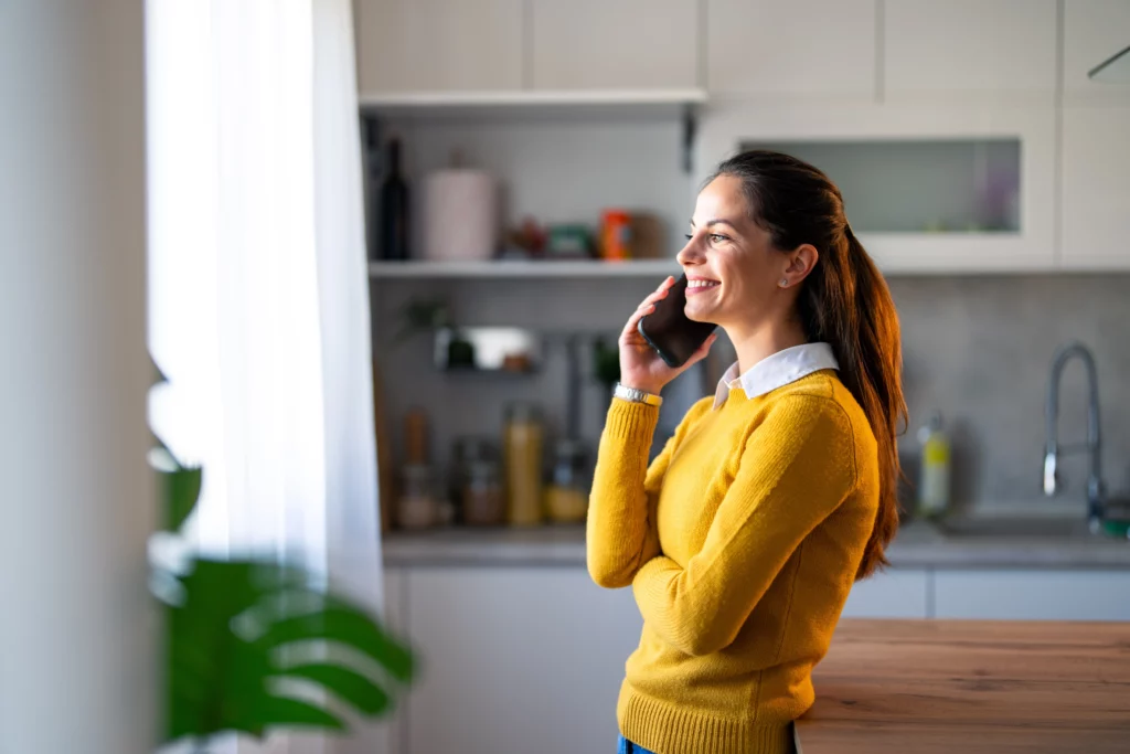 A woman in a yellow jumper smiles while taking a phone call in her kitchen