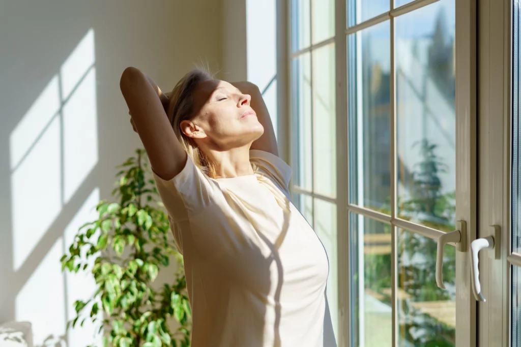A woman with her eyes closed and arms above her head stretches while soaking in the morning sunlight from her window