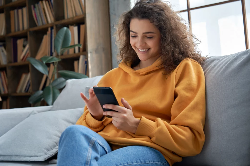 A woman smiles at her phone while sitting on a grey couch