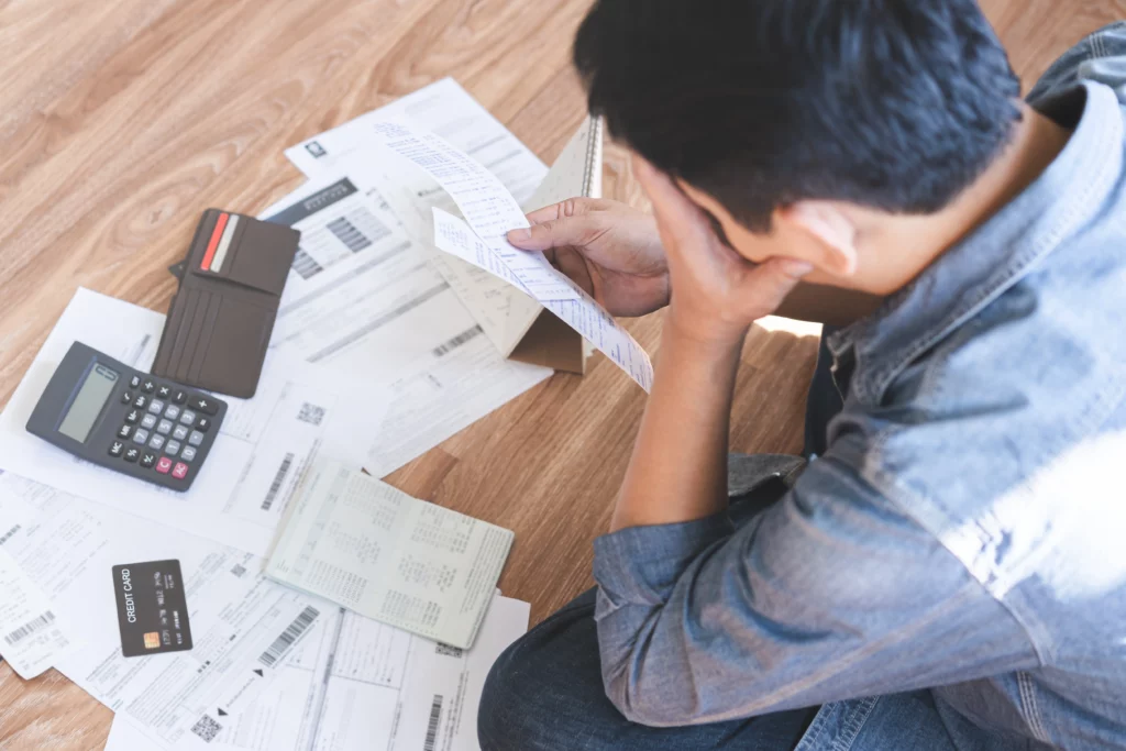 A man sits on the floor in front of a pile of unpaid bills, his empty wallet, and a calculator displaying zero