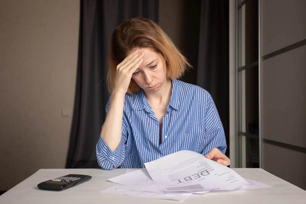 A stressed woman with a hand on her forehead looks at a pile of bills and debt